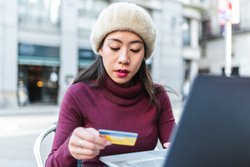Concentrated Chinese female with credit card making online purchase on laptop while sitting on terrace of cafe in modern city