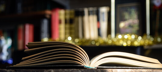 The open book, illuminated by the sun, lies on the table. Bookshelf background and blurry color spots.