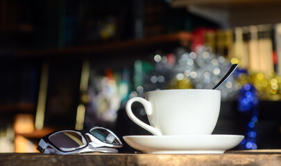 A sunlit coffee cup against the backdrop of bookshelves. There are glasses next to it.