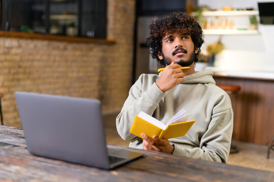 Male student pondering and looking away while writing lecture in a notebook at home. He using laptop to watching a lecture
