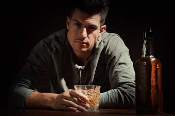 Addicted man with glass of alcoholic drink at wooden table against dark background