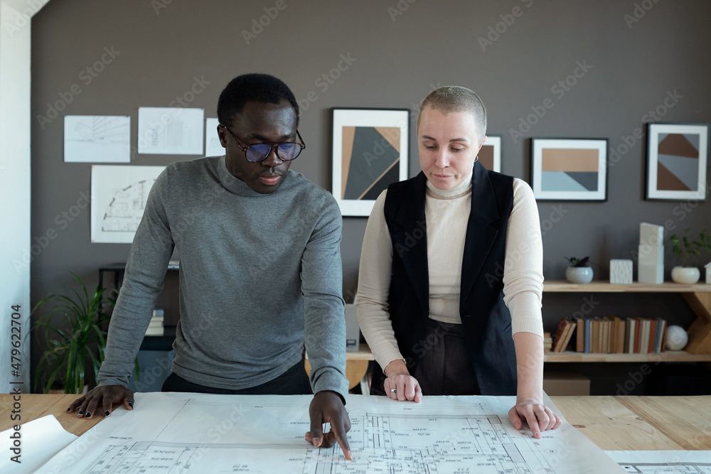 Poster Contemporary creative architects discussing sketch on blueprint while one of them pointing at size notes