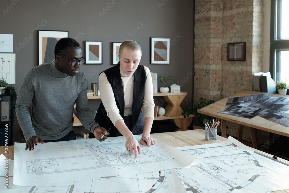 Poster Two confident engineers in smart casualwear discussing sketch on blueprint while standing by table in large office