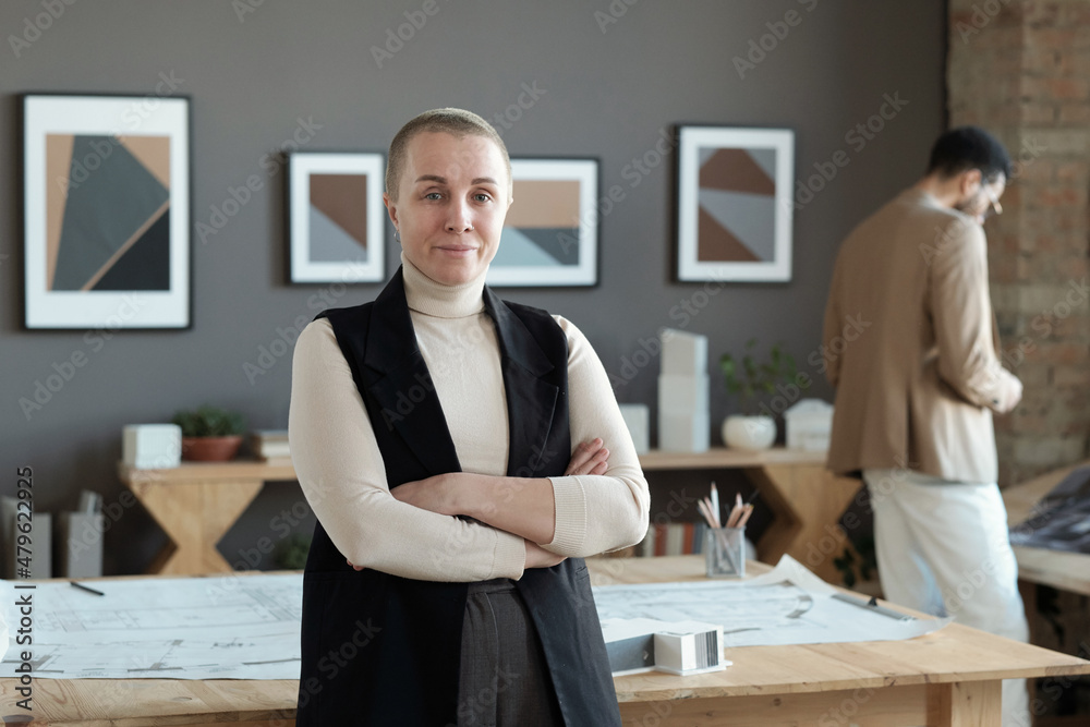 Poster successful female architect crossing arms by chest while standing by table with blueprints on backgr