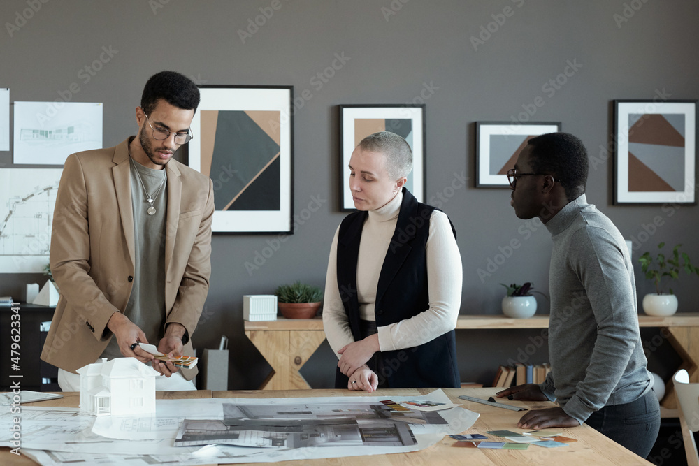 Poster Group of intercultural colleagues choosing color from palette while one of them holding swatches over blueprints and sketches