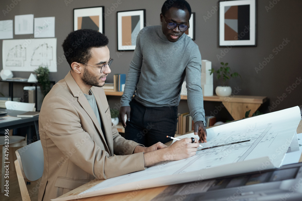 Poster Two young intercultural businessmen discussing sketch of architecture on blueprint while one of them drawing lines