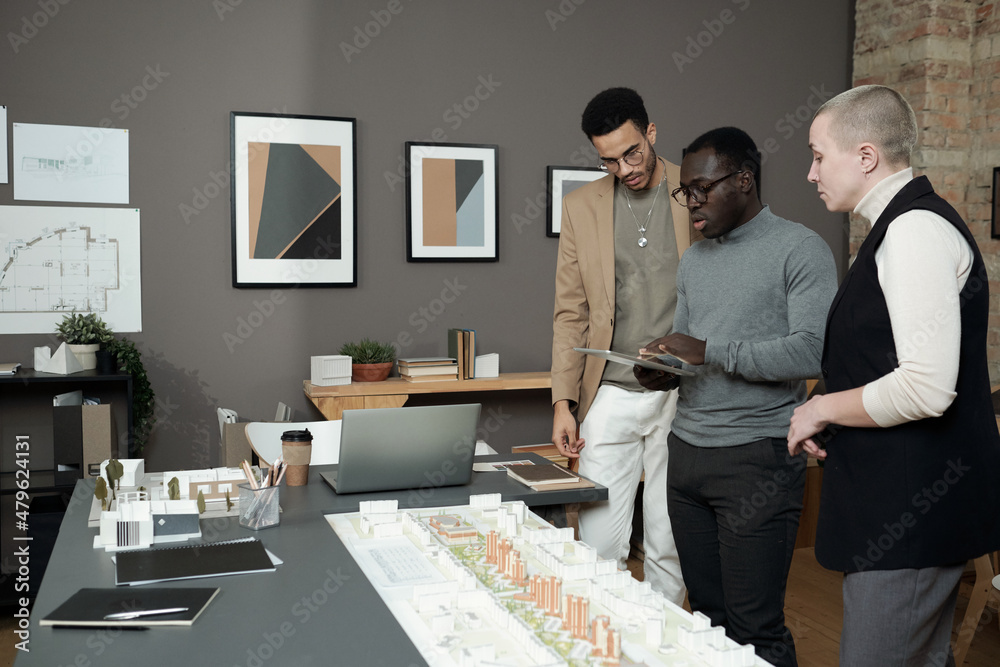 Poster Three creative designers or architects discussing layout at meeting while African man with tablet making presentation
