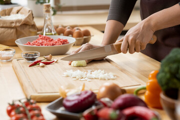 Hands of young female chopping fresh onion on wooden board while preparing italian pasta with...