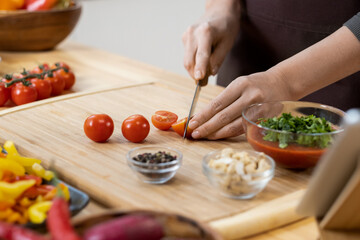 Hands of young female chopping fresh ripe cherry tomatoes for salad on wooden board while standing by kitchen table