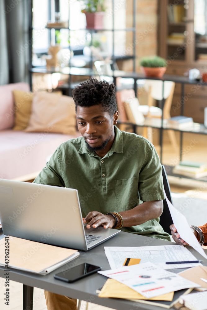 Poster young businessman of african ethnicity looking at laptop display while networking by table in home e