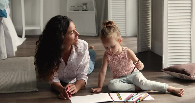 Latina woman cute 4s girl drawing with colored pencils, lying on warm wooden floor in cozy bedroom create paintings in album enjoy hobby and communication. Mother and daughter pastime at home concept