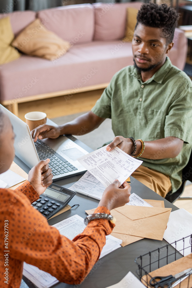 Poster young african man passing financial paper to female while sitting by table in front of laptop and wo