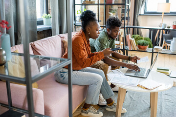 Two young contemporary coworkers working with mobile gadgets and financial data while sitting on couch by table