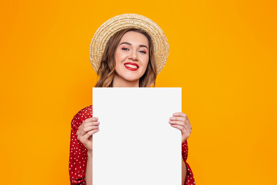Portrait of a young woman in a red dress, a straw hat with red lips smiling and holding in her hands an A3 format poster isolated over orange background, mockup