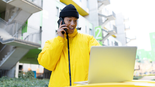 Cheerful Asian man chatting on smartphone while browsing laptop