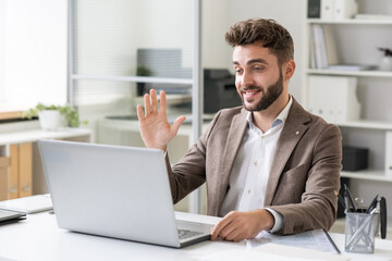 Happy young businessman waving hand to someone on laptop screen while communicating during online meeting