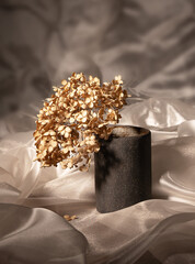 A ceramic glass with a dried hydrangea flower on a background of flowing, silk fabric. Minimalism, monochrome composition. Interior design concept.