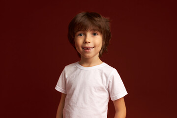 Indoor close-up portrait of adorable curious male child with sincere vivid smile dressed in white isolated over red wall background, listening with enthusiasm to his interlocutor. Carefree childhood