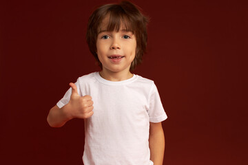 Happy excited little boy in white showing excellent and perfect symbol with hand, putting thumb up, standing against red background, having cute sweet face and big blue eyes. Carefree childhood