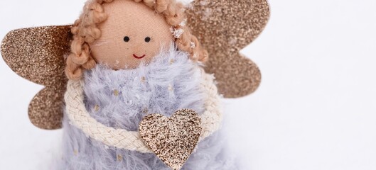 A soft textile doll with golden heart in hands sitting on the snow. Valentine’s Day, love, Merry Christmas, New Year concept. Banner.