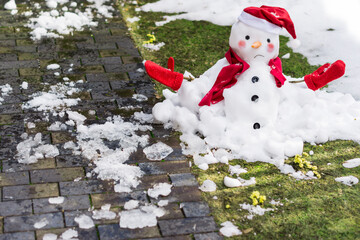 Unhappy snowman in mittens, red scarf and cap is melting  outdoors in sunlight on snowy green grass...