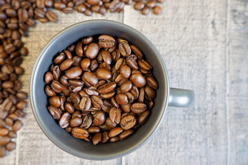 a dark gray mug filled with roasted whole coffee beans stands on a wooden background with coffee beans on the left