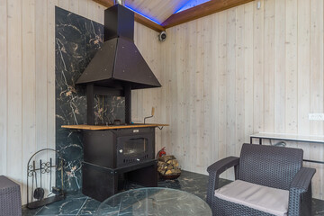 A room in a house with light wooden walls and granite floor tiles. Rattan furniture and cast-iron fireplace.