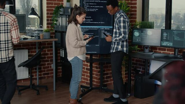 Team of database admins analyzing source code on wall screen tv comparing errors using digital tablet in busy server room. Two cloud programers debugging algorithm in software innovation office.
