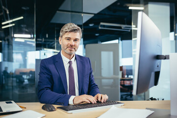 Portrait of a successful gray-haired man with a beard, businessman working at a computer in a modern office and looking at the camera