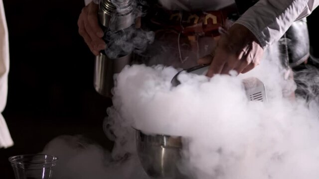 A man cooks ice cream. A man's hand pours liquid nitrogen into a cup and whips it with a mixer. Steam goes in different directions