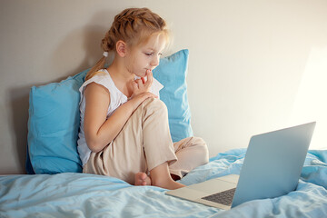 A cute girl sitting on the bed is watching something on a laptop. The concept of education