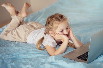 A cute girl sitting on the bed is watching something on a laptop. The concept of education