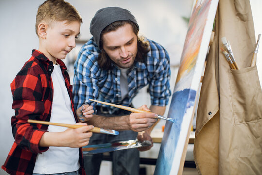 Focused caucasian man with paint brush in hands teaching little boy drawing on canvas at modern school. Teacher and student creating masterpiece together.