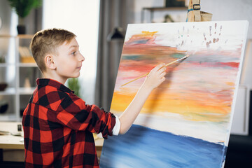 Side view of focused caucasian boy in casual outfit using brush for painting on easel at sudio. Favorite hobby, inspiration and talent concept.