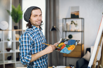 Portrait of talented young man in stylish casual wear holding color palette and paintbrush in hands while standing in front of easel. Concept of creativity and inspiration.