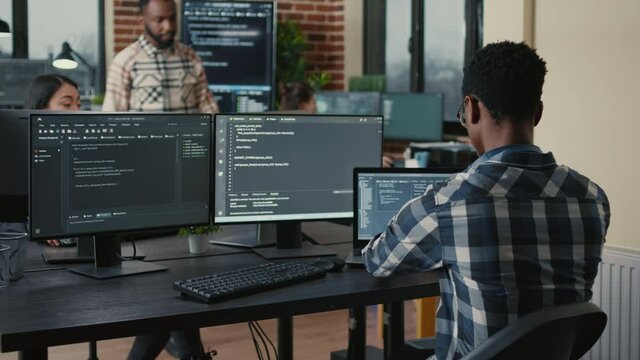 Software developer writing code on laptop and computer keyboard looking at multiple screens with programming language is interrupted by coworker asking for advice. Cloud programers doing teamwork.