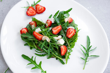 Romantic salad of strawberry with mix of herbs, arugula, spinach leaves, feta cheese and cashew nuts in heart shape on white plate. Romantic meal for St Valentine Day celebration.