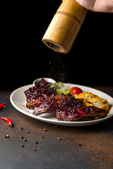 fried pork ribs with lingonberry sauce with pepper is poured from the pepper mill on dark brown table