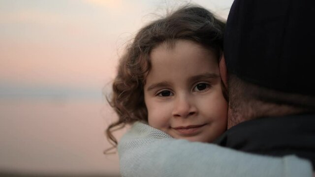 Portrait of a girl looking at the camera and hugging her grandfather with sunset in the background. Grandfather holds his granddaughter in his arms. Carefree quiet retirement concept.