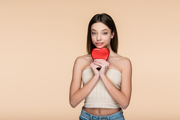 brunette young woman in crop top holding red heart-shaped metallic box isolated on beige