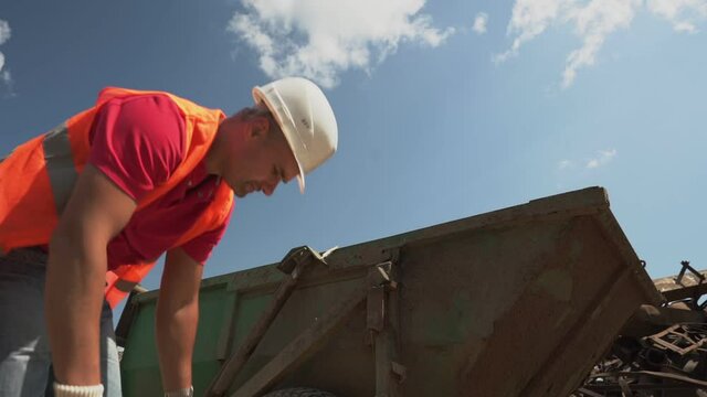 worker in helmet and retroreflective vest tilts downward collects wooden debris and loads into container outdoors closeup