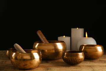 Obraz na płótnie Canvas Golden singing bowls with mallets and burning candles on wooden table against dark background