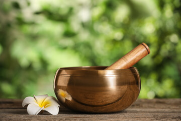 Golden singing bowl, mallet and flower on wooden table outdoors