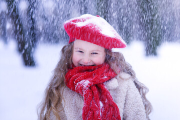 Portrait of happy positive cheerful joyful little girl, cute kid. Beautiful child is walking at winter park at snowy cold frosty day in red hat and scarf smiling, having fun. Snowfall, blizzard