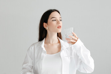 Studio portrait of young brunette woman in a white shirt and top, drinking glass of pure water, looking to the side, on white background. Purity concept