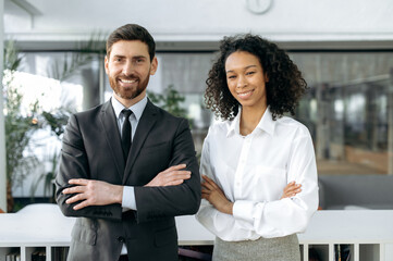 Influential successful coworkers of different nationalities, caucasian man and african american woman, in formal clothes, are standing with arms crossed in modern office, looking at camera and smiling