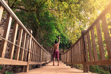 Fototapeta na wymiar Hombre joven latino de espaldas, levantando un brazo, parado en un puente de madera en medio de la selva