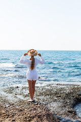 Fototapeta na wymiar An attractive young woman with long blond hair in a white summer suit stands on the seashore and enjoys the view of the ocean.