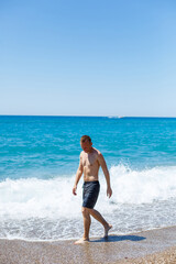 Fototapeta premium Attractive young man with a beautiful body in shorts walks along the sandy seashore and enjoying summer vacation