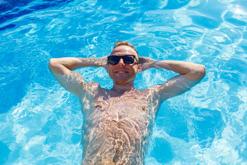 Fototapeta na wymiar Young attractive man in sunglasses is resting in the pool on summer vacation. The guy in the pool by the hotel